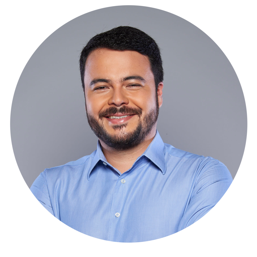 Foto de Rodrigo sorrindo, de braços cruzados. Ele é um homem branco, tem cabelo curto, barba cheia e veste camisa social azul-clara.