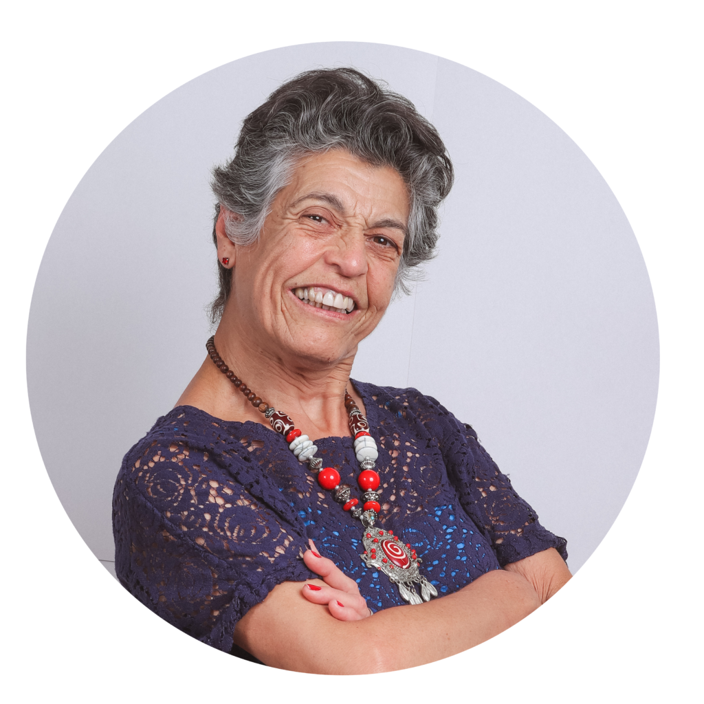 Mulher de pele clara e cabelos curtos, grisalhos, penteados para trás. Está sorrindo, com os braços cruzados. Veste blusa de renda azul e usa colar grande com contas vermelhas, brancas e detalhes prateados, com um pingente central decorado.
