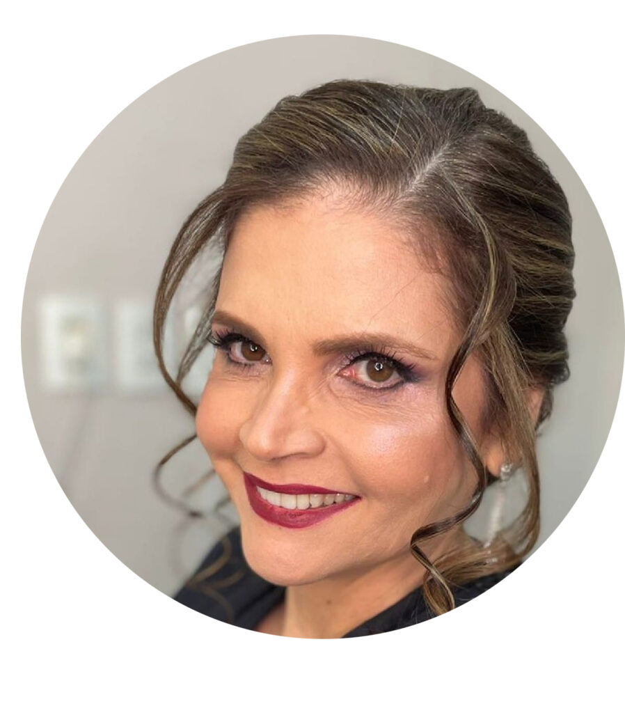 Fotografia colorida, em close, de uma mulher com pele clara, cabelos castanhos com mechas claras, penteados para trás em um coque, com algumas mechas soltas e onduladas. Ela tem olhos castanhos. Usa brinco pequeno e brilhante.
