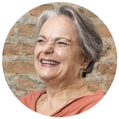 Mulher de pele clara, com cabelos curtos grisalhos, sorri com a cabeça levemente voltada para a esquerda. Veste blusa em tom salmão e brincos escuros. Ao fundo, há uma parede de tijolos aparentes.