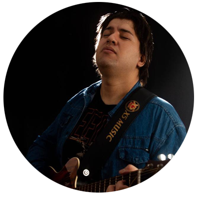 Sim, fica mais preciso mencionar a peça como camisa jeans. **Audiodescrição:** Homem de pele clara, com cabelos escuros e médios, aparece tocando violão. Está com os olhos fechados e a cabeça levemente inclinada para trás, em uma expressão de concentração. Veste camiseta preta e camisa jeans azul. A correia do violão cruza o ombro e parte do instrumento aparece na região inferior da imagem. O fundo é escuro, com iluminação direcionada sobre a pessoa.