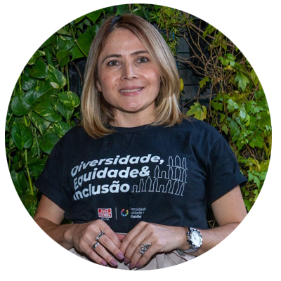 Mulher adulta de pele clara, com cabelos loiros na altura dos ombros, lisos e repartidos de lado. Está voltada para a câmera, sorrindo suavemente. Usa camiseta preta com a frase “Diversidade, Equidade & Inclusão” em destaque na parte frontal. Também usa relógio, anéis e esmalte claro nas unhas. Ao fundo, há uma parede coberta por folhagens verdes. A imagem mostra a pessoa da cintura para cima.
