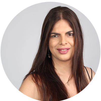 Mulher trans de pele clara, com cabelos longos, lisos e castanho-escuros, repartidos ao meio. Tem sobrancelhas espessas e escuras, olhos castanhos, usa maquiagem discreta com batom rosa e sorri suavemente para a câmera. Veste uma blusa preta de alças finas.