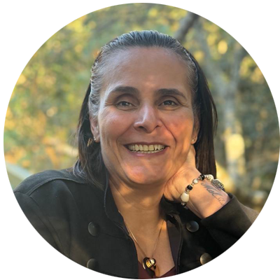 Mulher sorridente, com cabelos escuros presos para trás, veste roupa preta e apoia o rosto sobre uma das mãos. Ao fundo, vegetação desfocada.