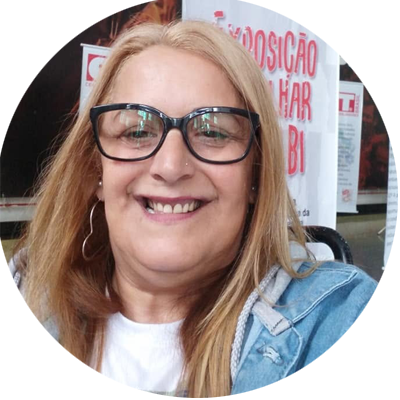 Mulher de pele clara, com cabelos longos, lisos e loiros, sorrindo para a câmera. Usa óculos de armação preta grande, brincos de argola, camiseta branca e jaqueta jeans. Ao fundo, há um painel com textos em vermelho e branco, desfocado.