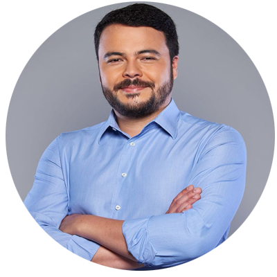 Homem de cabelos curtos e escuros, barba, vestindo camisa azul-clara de mangas compridas. Está de braços cruzados, diante de fundo cinza, em foto recortada em formato circular.